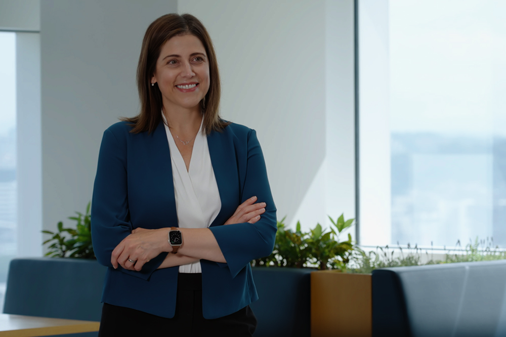 Danielle Merfeld, standing with arms crossed and smiling in an office with large windows and plants in the background.